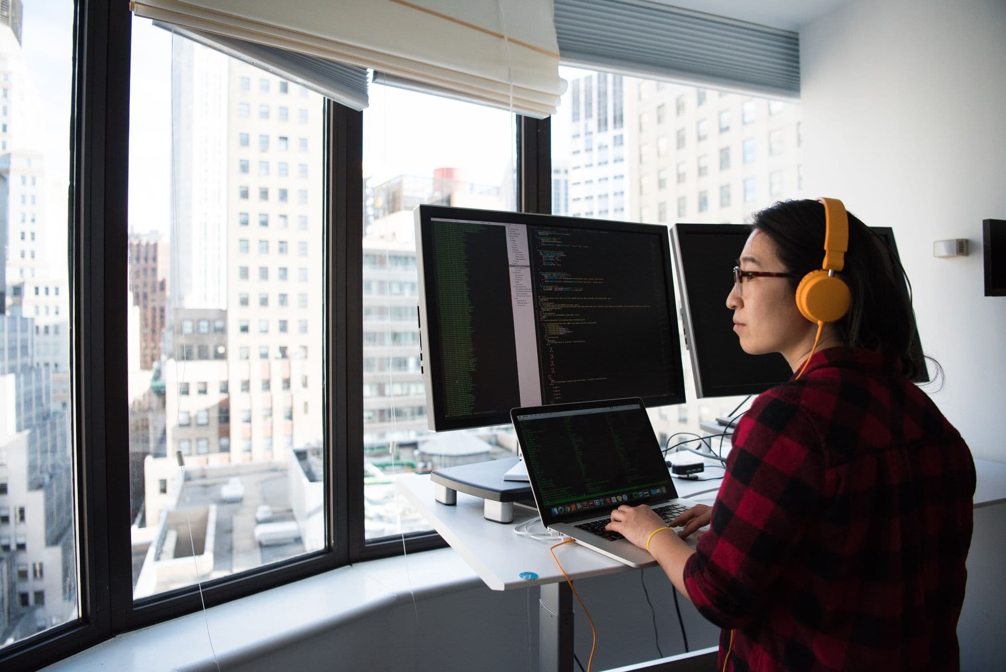 A person working alone at a desk in a dark room, headphones on, two monitors lit, a city window behind them.