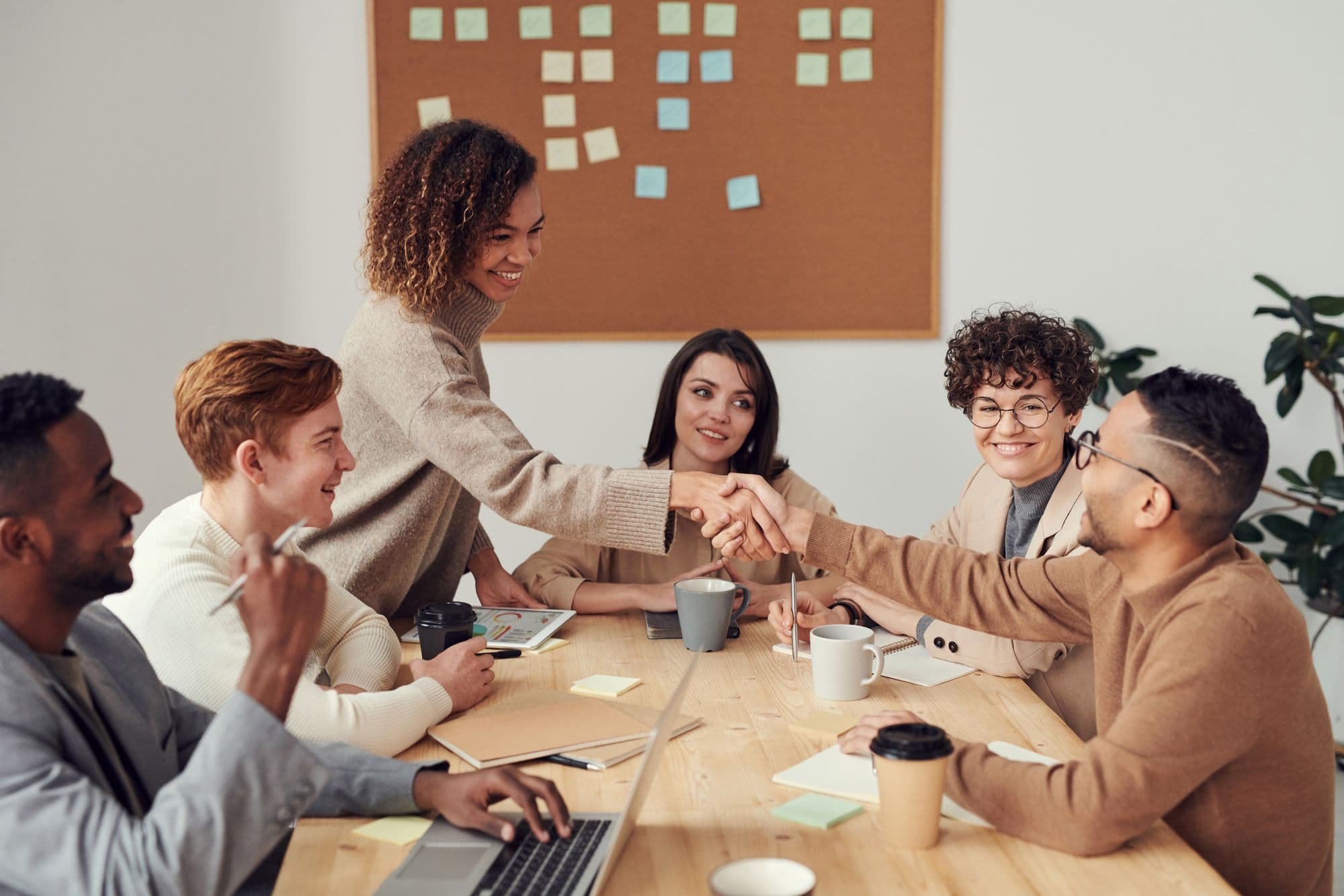 A meeting around a table, five people mid-conversation, two of them shaking hands, daylight from a window behind.