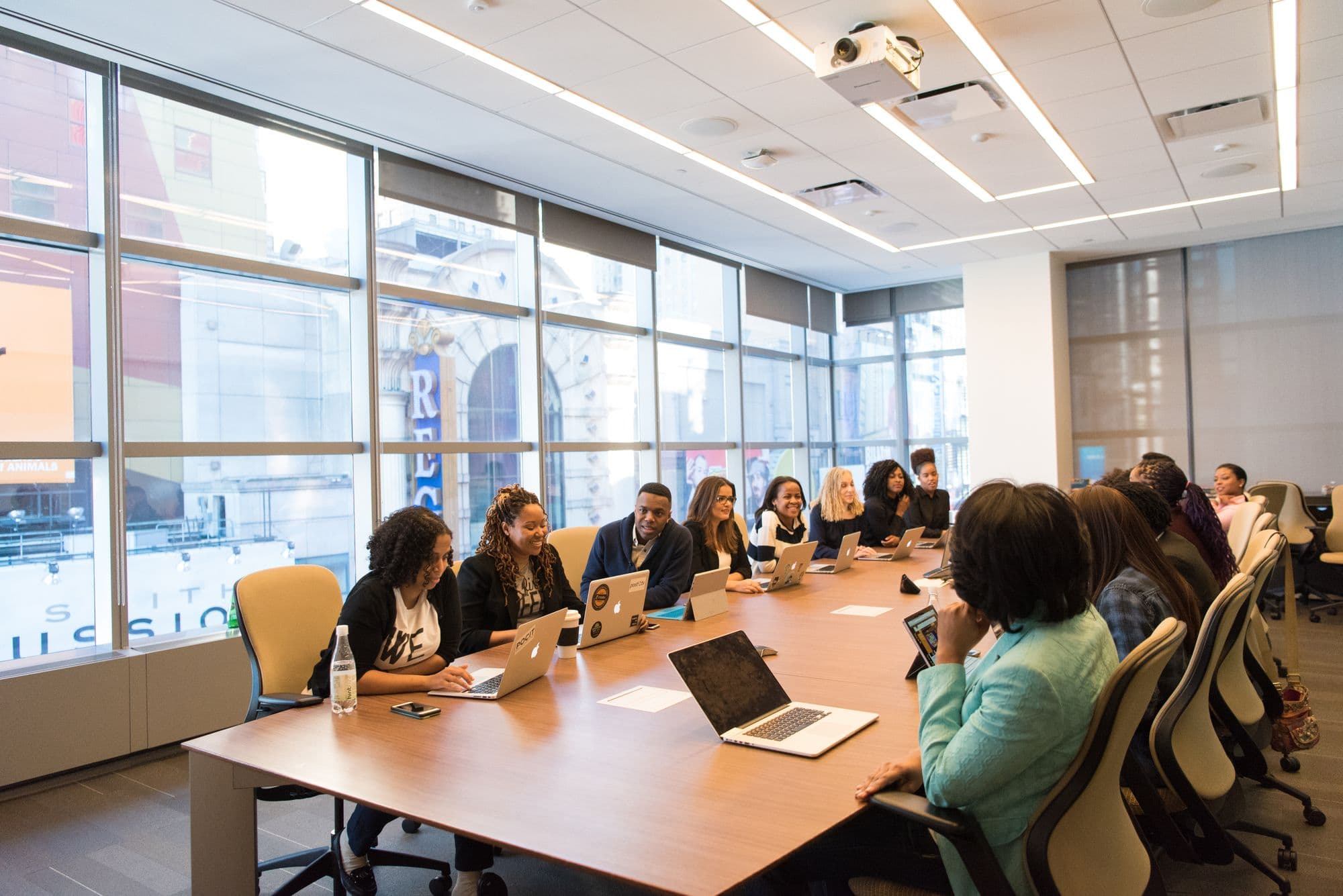 A wide conference room seen from the back, a long table of people facing a lit screen, tall windows down one side.